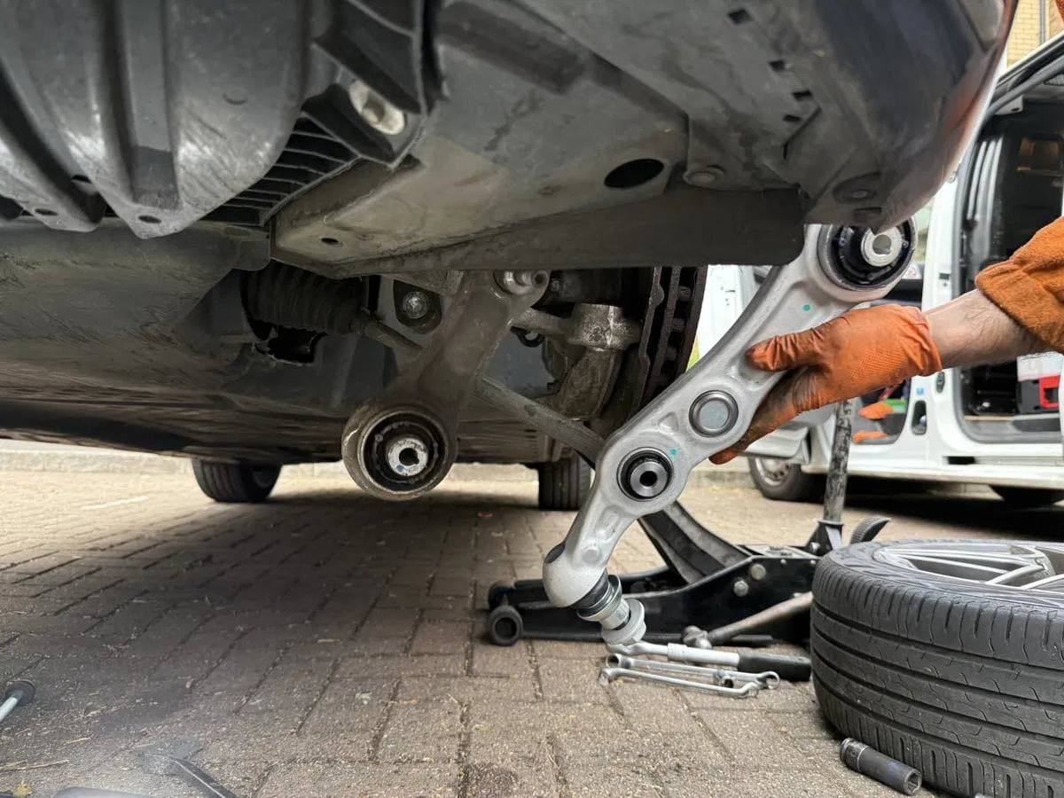 Mobile mechanic replacing a front suspension control arm at the roadside, van parked alongside